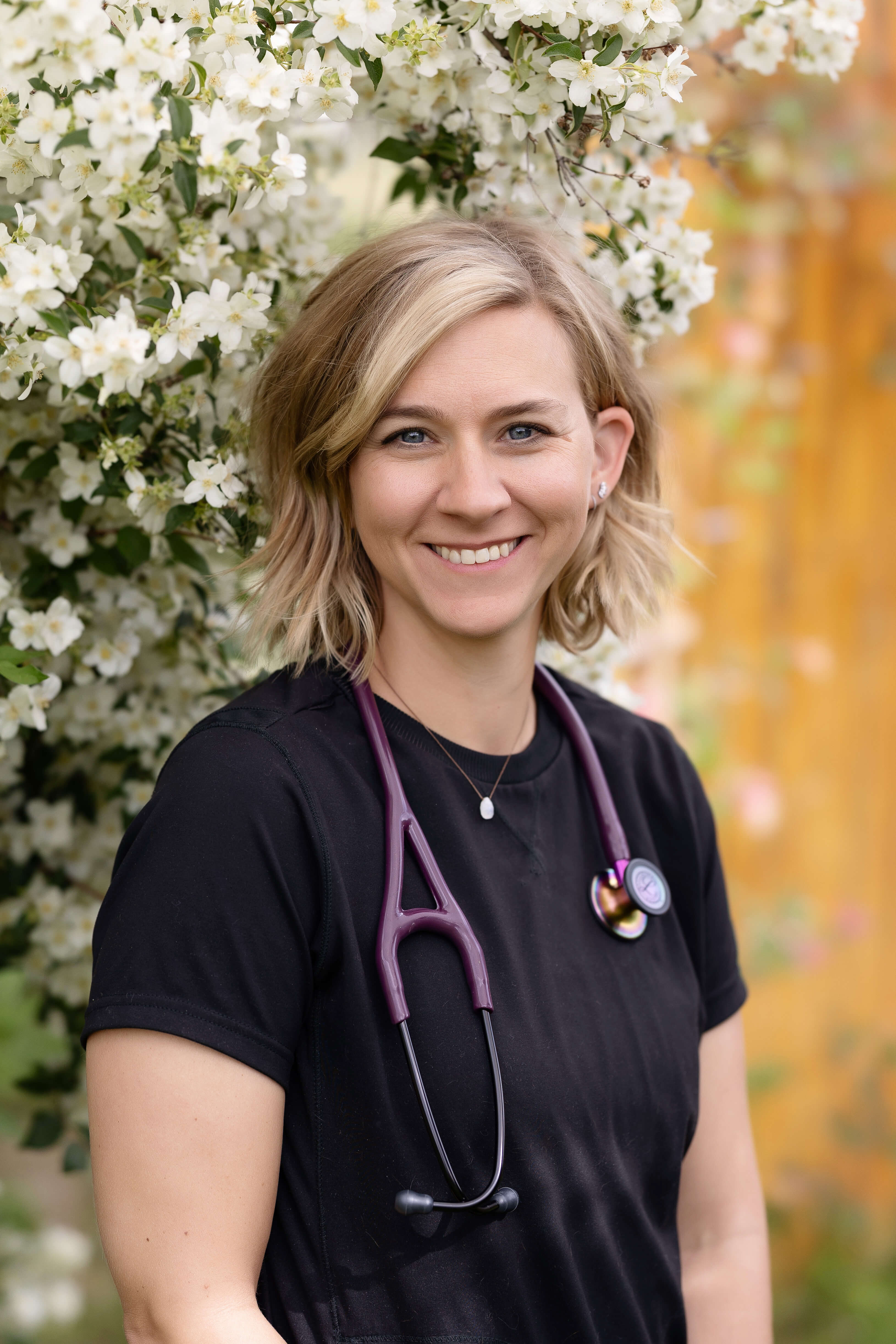 Blonde woman in black scrubs, with stethoscope in front of a flowing shrub.