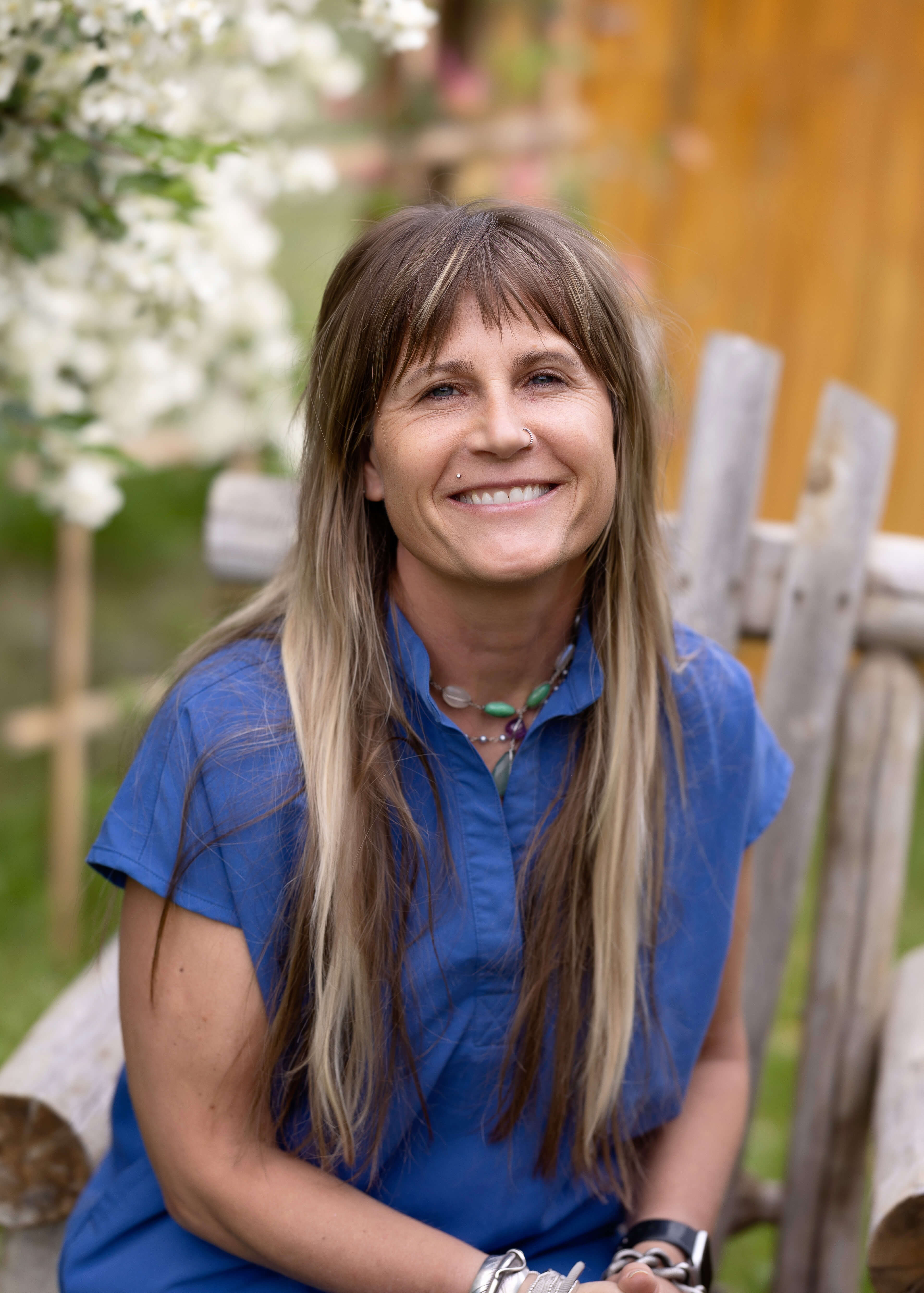 Woman with long hair, in blue scrubs, sitting in a wooden chair outside.
