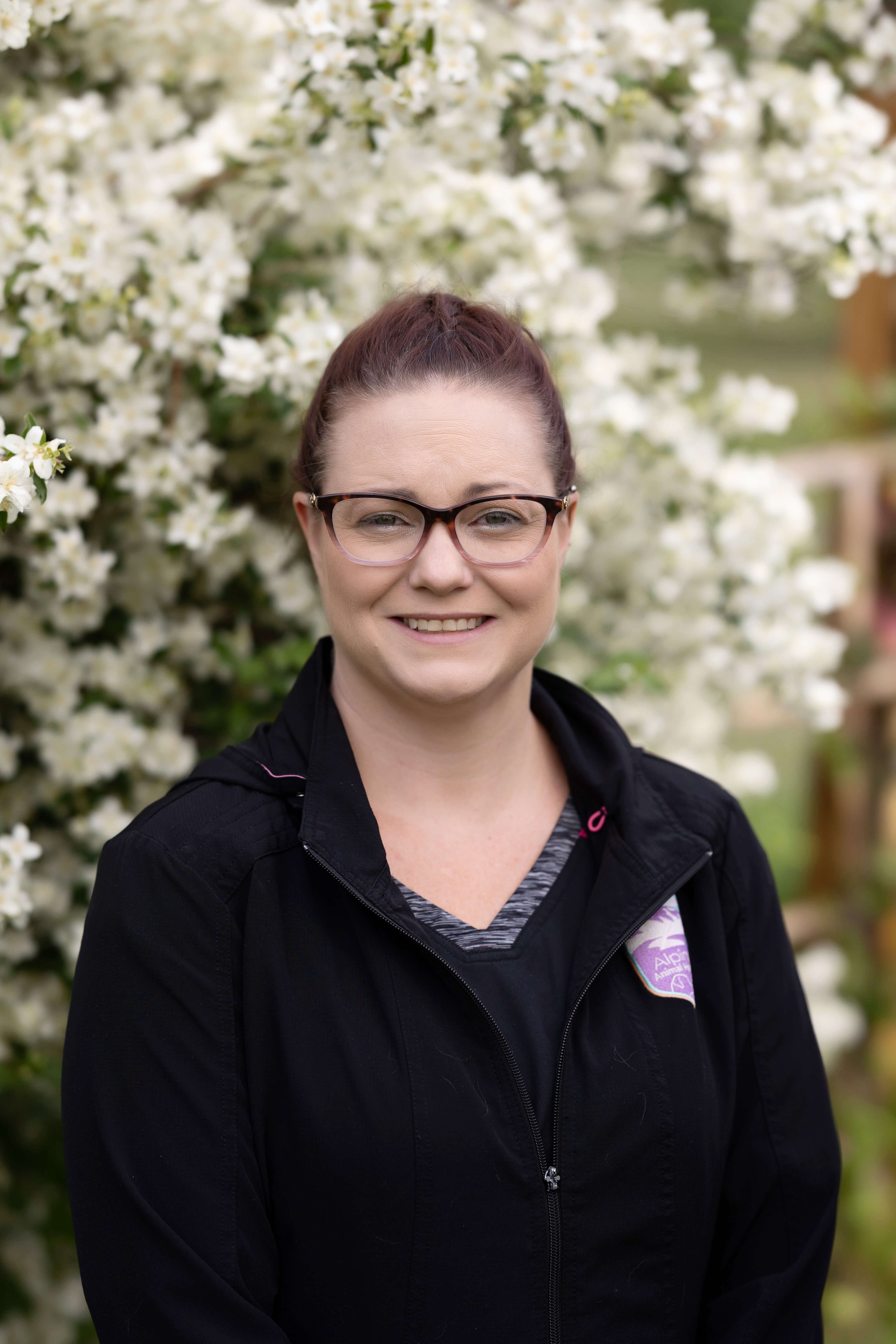 Brunette in black scrubs, wearing glasses, outside in front of a flowering shrub.