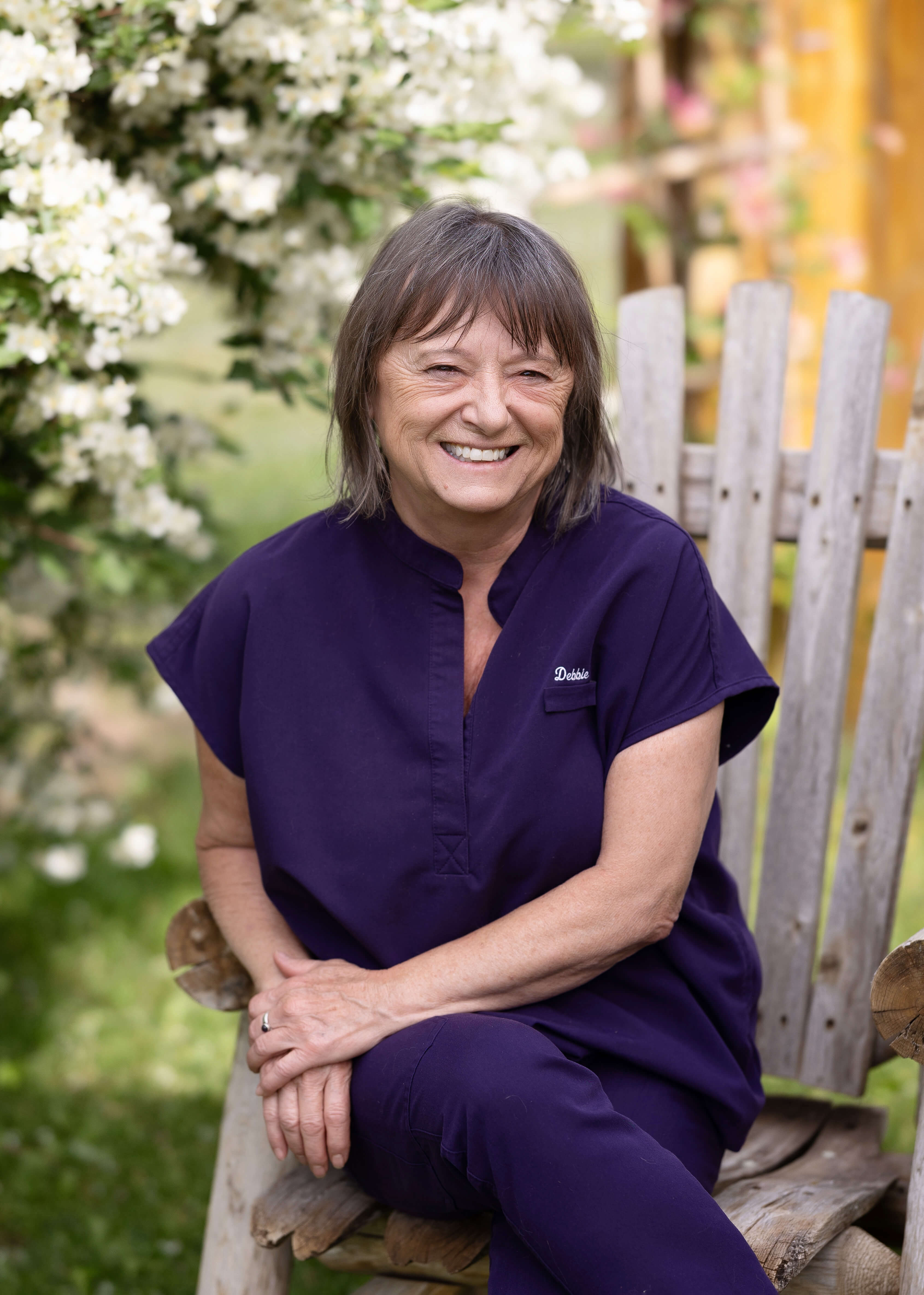 Smiling woman in purple scrubs, sitting in a wooden chair outside.