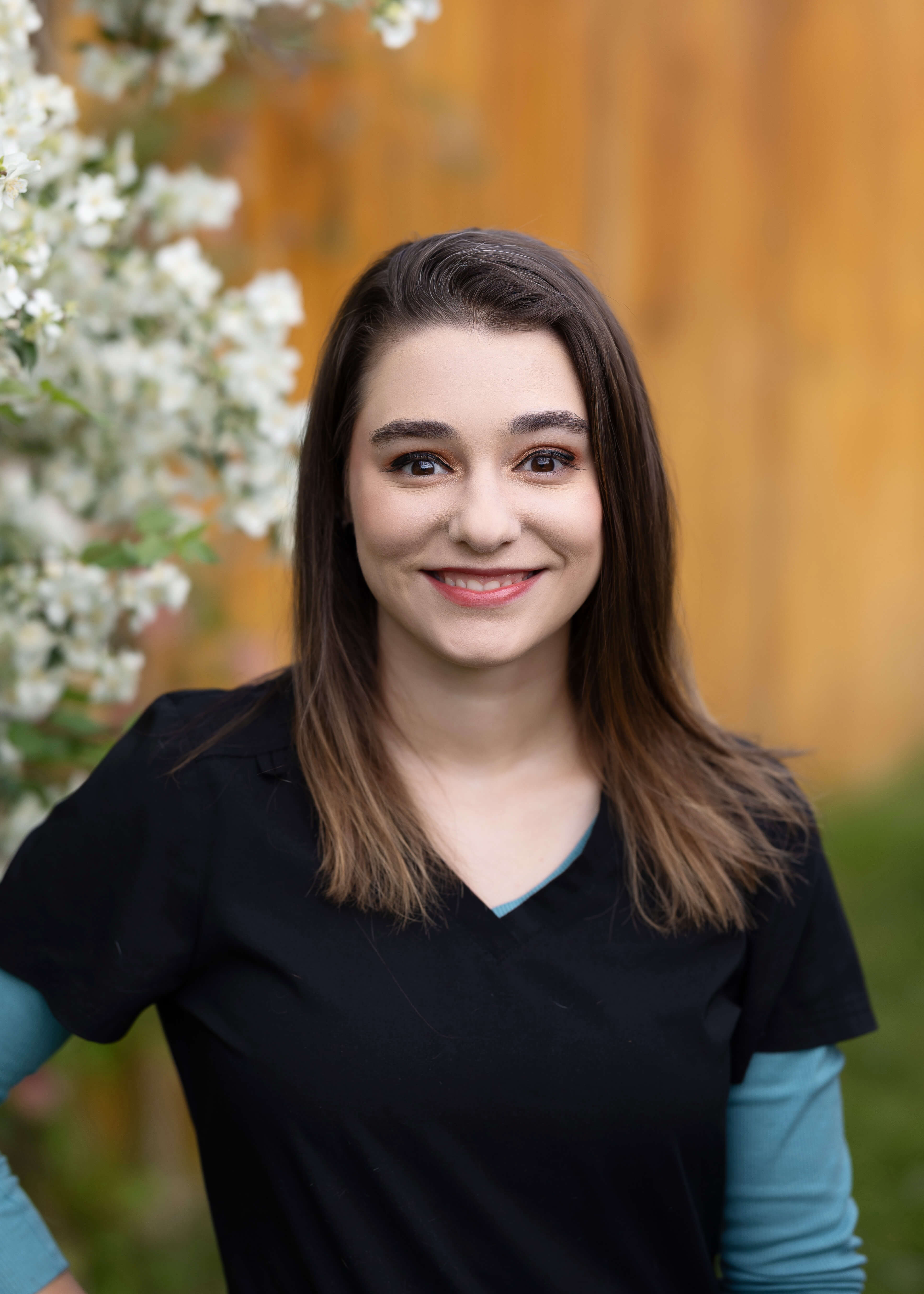 Brunette in black scrubs outside in front of a flowering shrub.