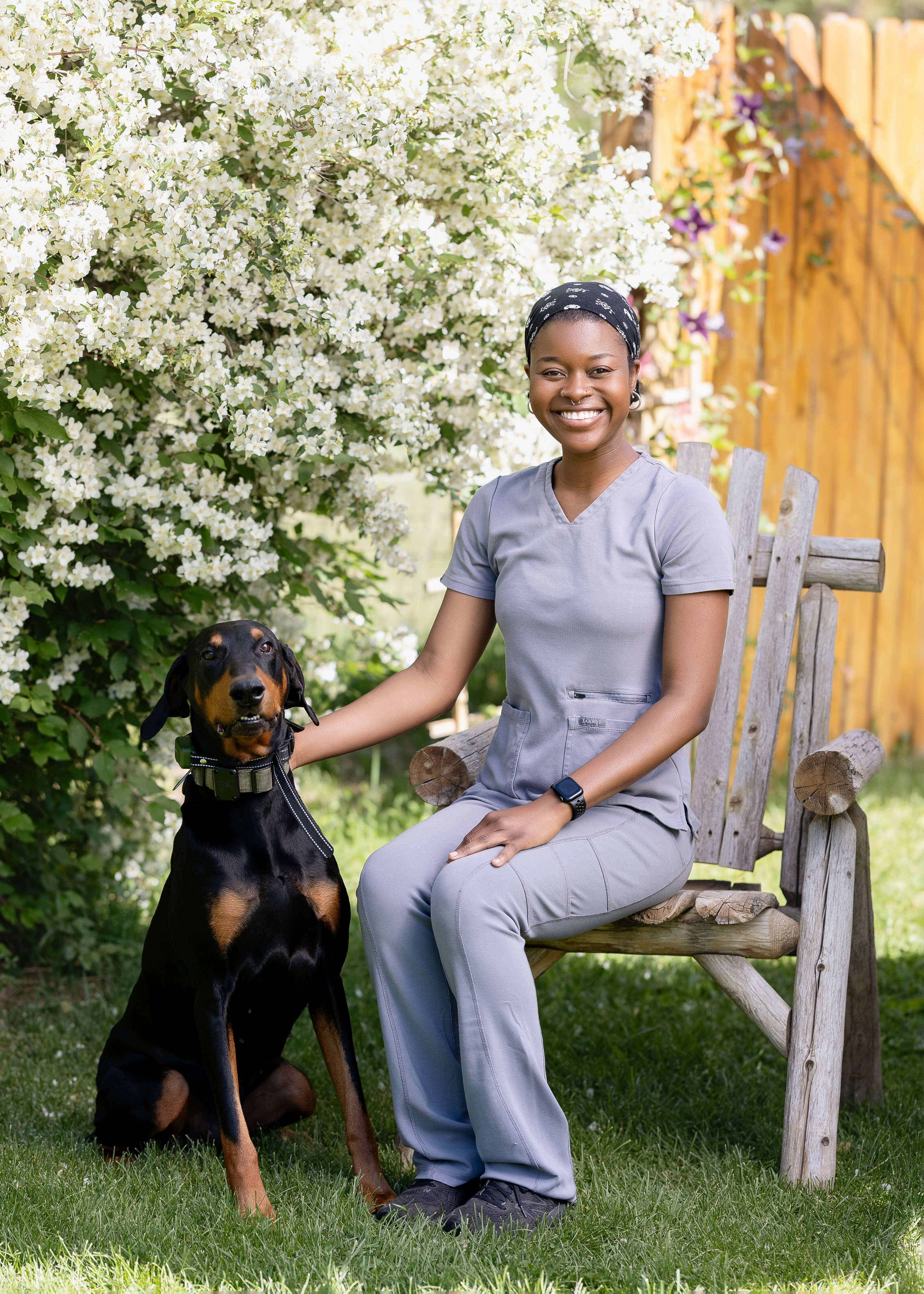 Woman sitting next to a doberman, outside in front of a flowering shrub.
