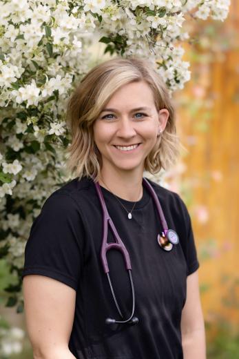 Blonde woman in black scrubs, with stethoscope in front of a flowing shrub.