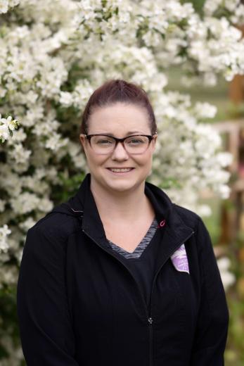 Brunette in black scrubs, wearing glasses, outside in front of a flowering shrub.
