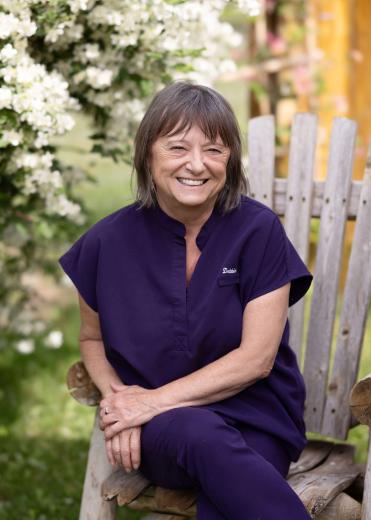 Smiling woman in purple scrubs, sitting in a wooden chair outside.