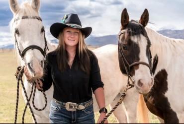A blonde woman in a black cowboy hat, standing between and holding two horses.
